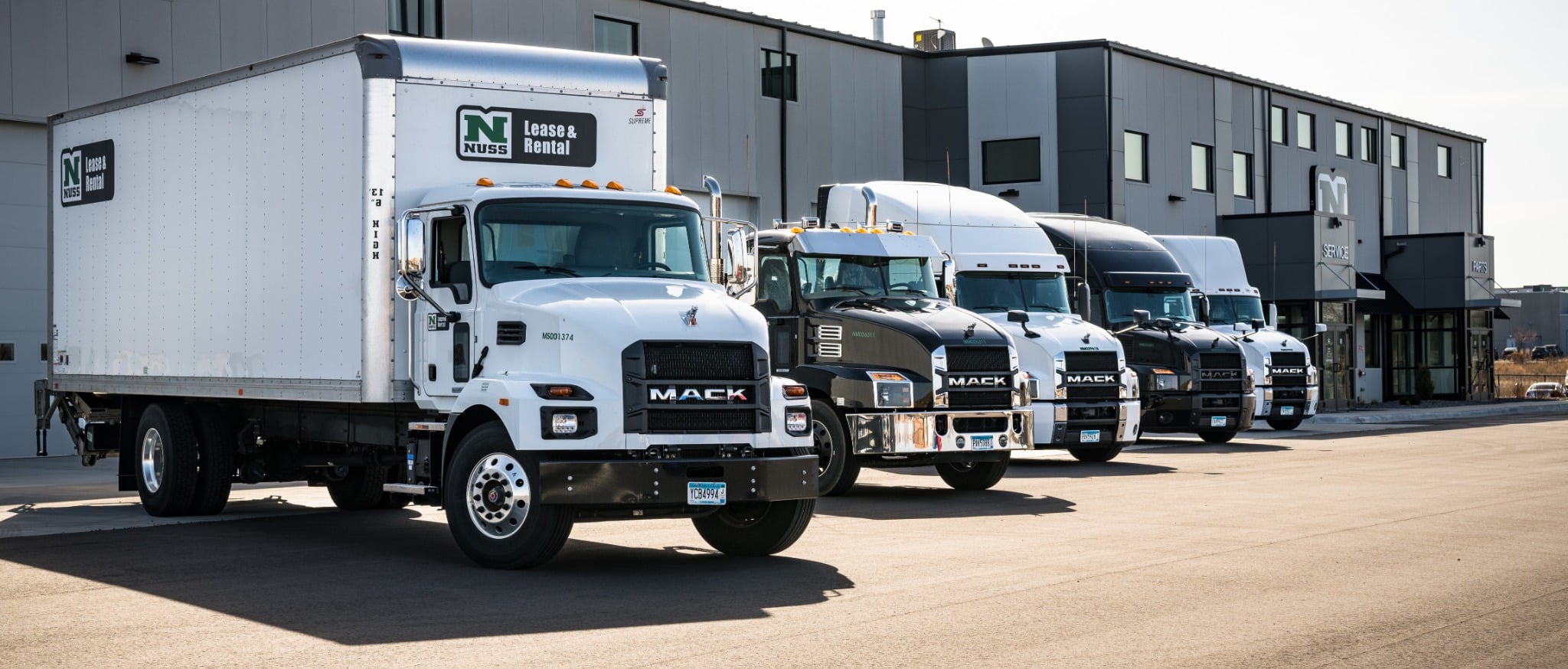 Nuss Lease & Rental Mack box truck in front of a lineup of Mack tractors at a Nuss facility